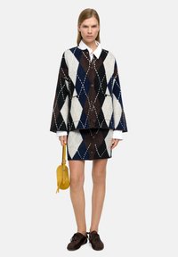 Sweter o większym rozmiarze z wzorem argyle w kolorach ciemno-brązowym, granatowym i kremowym, w połączeniu z pasującą spódnicą; posiada guziki i wyrazisty biały kołnierz.