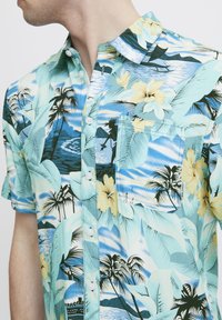 Camicia a manica corta con stampa floreale tropicale e palme in turchese, giallo e verde. Presenta una chiusura con bottoni e una tasca sul petto.