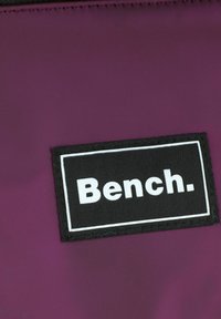 Tessuto di nylon viola con un'etichetta rettangolare nera che presenta il testo bianco "Bench." in un carattere audace e moderno. Texture liscia, design minimalista.