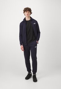 Marineblå joggingdräkt tillverkad av mjukt tyg, med en zip-jacka med hög krage och matchande avsmalnande byxor, båda med logotypdetaljer.