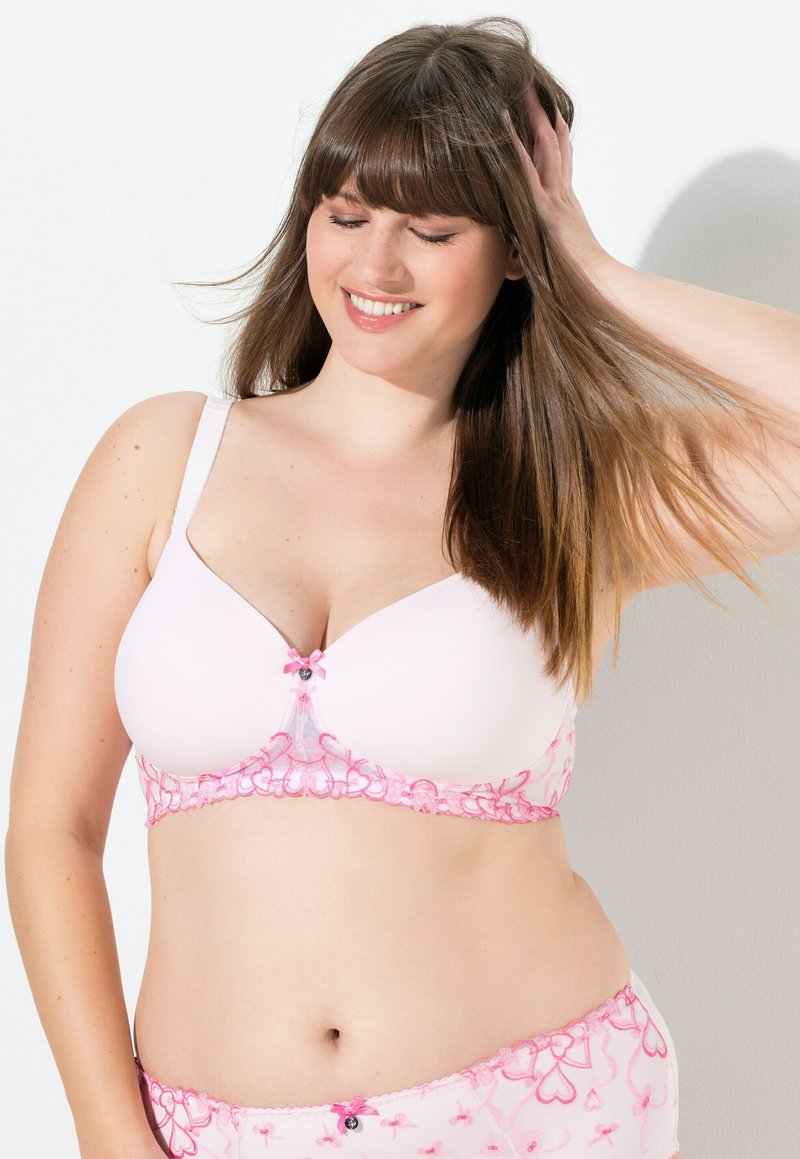 Ljust rosa bh med spetskant i blommönster, prydd med en liten svart och rosa rosett i mitten. Slät textur, mjukt tyg.
