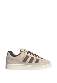 Beige suède sneakers met bruine accenten, voorzien van drie strepen, een getextureerde zool en een ronde neus. Vetersluiting en een logo aan de zijkant.