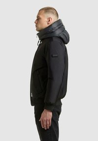 Schwarze gesteppte Jacke mit hohem, voluminösem Kapuzenkragen, Reißverschlusstasche vorne, elastischen Bündchen und strukturiertem Stoff. Schmale Passform mit modernem Design.