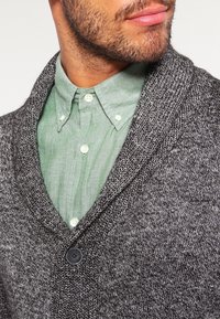 Gray knit cardigan with a textured finish, featuring a wide shawl collar and a single button closure over a light green button-up shirt.