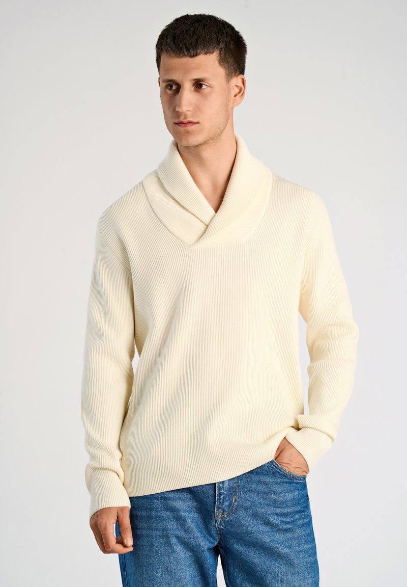 Cremfarbener gerippter Pullover mit Schalkragen, langen Ärmeln und lockerer Passform, kombiniert mit blauen Jeans.