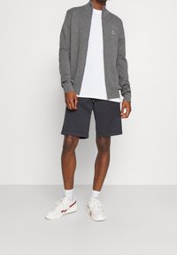 Cárdigan gris con cremallera, camiseta blanca debajo y pantalones cortos negros. Zapatillas blancas con detalles rojos. Textura suave, ajuste casual.
