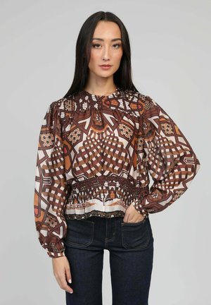 Jeune femme aux longs cheveux foncés portant un chemisier marron à motifs géométriques avec des manches bouffantes et un jean foncé, debout avec une main dans la poche.