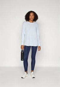 Top de manga comprida azul claro combinado com leggings azul-marinho, com uma textura suave. A modelo segura uma garrafa de água preta e usa ténis brancos.