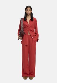 Kobieta stoi w czerwonej koszuli zapinanej na guziki i szerokich spodniach, trzyma ciemnobrązową skórzaną torbę na ramieniu, ma na sobie brązowe buty.