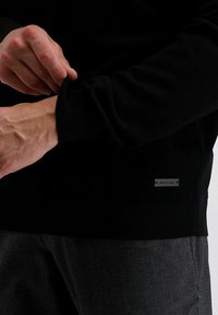 Schwarzer Pullover mit gerippten Bündchen, ausgestattet mit einem kleinen silbernen Logohangtag. Der Stoff wirkt glatt, hat einen figurbetonten Schnitt und strukturierte Ärmel.