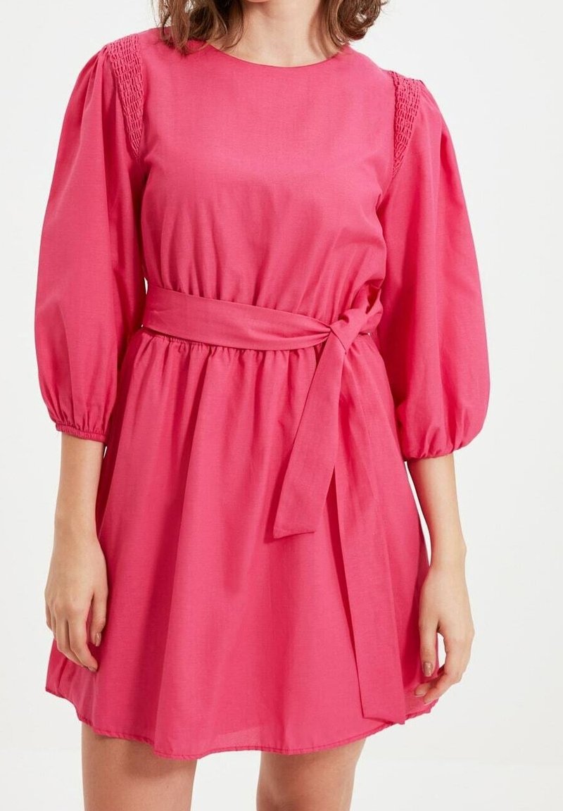 Femme portant une robe rose vif, arrivant aux genoux, avec des manches trois-quarts bouffantes et une ceinture nouée à la taille, debout devant un fond blanc.