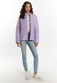 Giacca a vento viola chiaro con cuciture orizzontali, chiusura con zip e colletto alto. Indossata sopra una maglietta a righe bianche e grigie, abbinata a jeans blu.