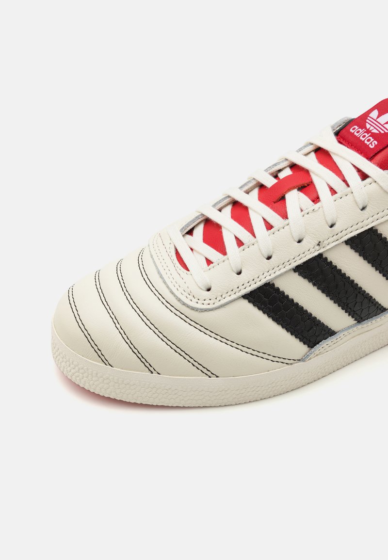 Sneaker bianco in pelle con strisce nere, dettagli rossi sulla linguetta e sui lacci, caratterizzata da dettagli testurizzati e una suola in gomma.