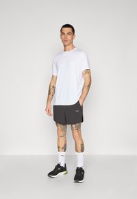 Hvid kortærmet t-shirt, sorte atletiske shorts, sorte sko med grønne detaljer, hvide sokker, viser forskellige tatoveringer på benene.