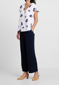 Blusa bianca con motivi astratti in navy, rosso e blu, scollatura a V, maniche corte, abbinata a pantaloni larghi navy e tacchi nudi.
