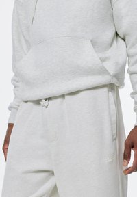 Lichtgrijze hoodie met een voorklep, gecombineerd met bijpassende joggingbroeken met een trekkoord en zijzakken. Subtiele branding op de broeken.