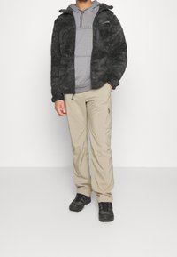 Giacca in pile nera e pelosa con chiusura a zip, felpa gray sotto, pantaloni cargo beige e scarpe da trekking nere. Design semplice e funzionale.
