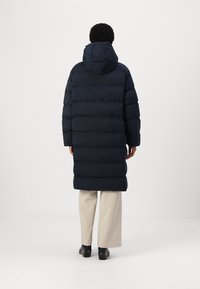 Navy puffer coat with a hood, featuring a quilted design, long sleeves, and a below-the-knee length. Worn over light-colored pants and footwear.