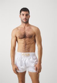 White satin boxing shorts with an elastic waist, featuring a "Venum" logo in gold. Model has a muscular build and short dark hair.