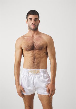 Hvit sateng bokse shorts med elastisk midje, med et "Venum"-logo i gull. Modellen har en muskuløs bygning og kort mørkt hår.