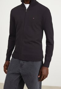 Czarny kardigan zapinany na zamek błyskawiczny z podniesionym kołnierzem, o dzianinowej fakturze, z prążkowanymi mankietami i dołem. Na piersi znajduje się małe logo.