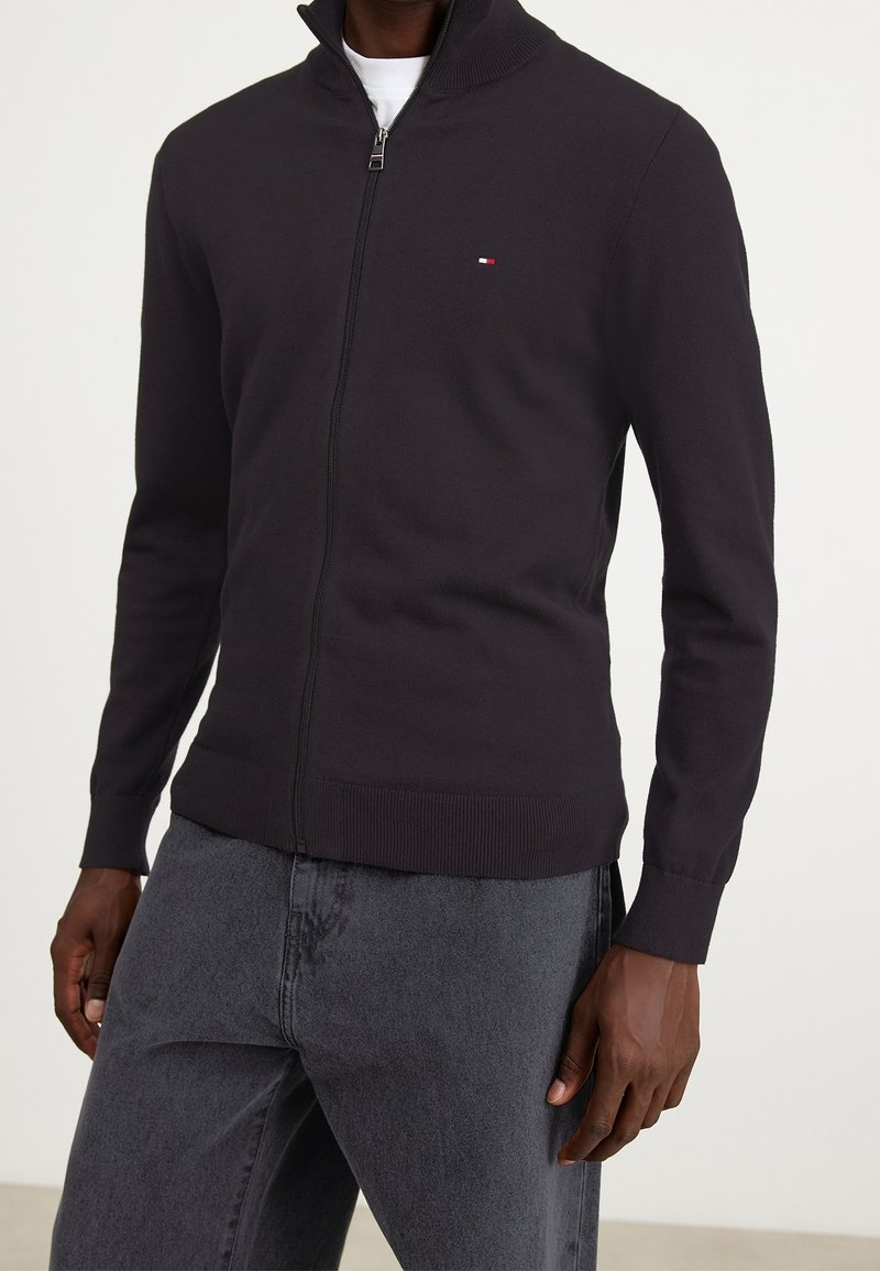 Czarny kardigan zapinany na zamek błyskawiczny z podniesionym kołnierzem, o dzianinowej fakturze, z prążkowanymi mankietami i dołem. Na piersi znajduje się małe logo.