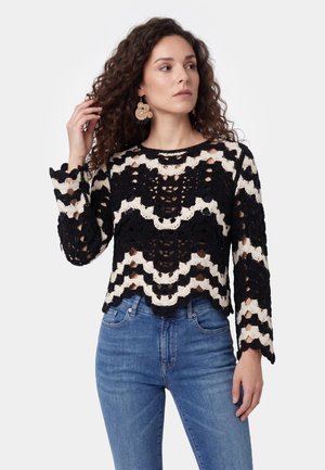 Kvinde med krøllet hår iført store runde øreringe, en sort-hvid langærmet top med hæklet mønster og blå jeans, der kigger til siden.