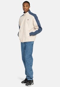Beige zip-up jacket with navy blue sleeves, elastic cuffs and waistband, paired with light blue jeans and black sneakers.