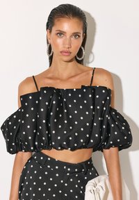 Femme aux cheveux lissés en arrière portant un top noir à pois et épaules dénudées avec un pantalon assorti, grandes boucles d’oreilles créoles, tenant une pochette blanche plissée.