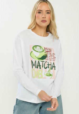 Blondynka w białej koszuli z długim rękawem z zielonym motywem matcha latte oraz napisem "Matcha Club" i wypisanymi pozycjami z menu.