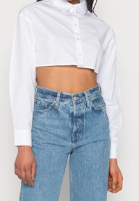 Camisa blanca de botones cortada con mangas largas, que presenta un cuello clásico y una textura suave, combinada con jeans de tiro alto de color azul claro.