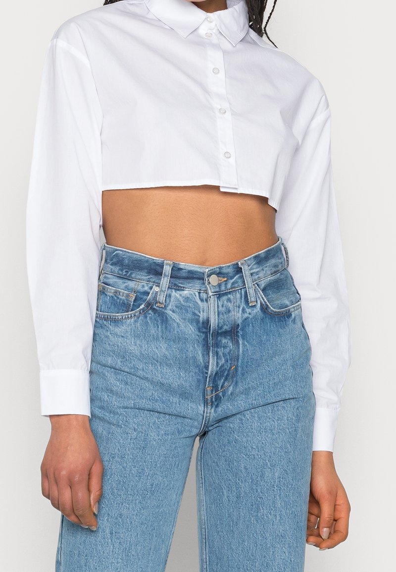 Camisa blanca de botones cortada con mangas largas, que presenta un cuello clásico y una textura suave, combinada con jeans de tiro alto de color azul claro.