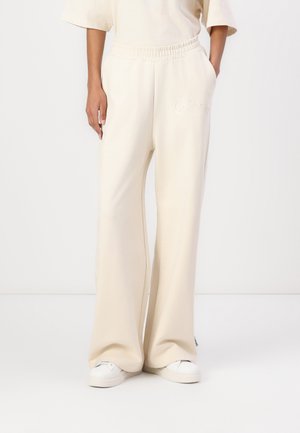 Kvinde iført cremefarvede sweatpants med vide ben og elastisk talje samt hvide sneakers, stående mod en ensfarvet baggrund.