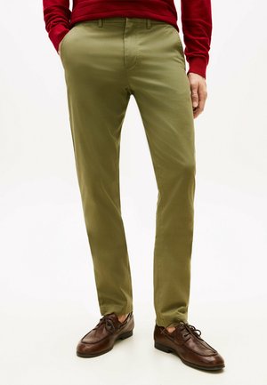 Mann trägt olivgrüne Chinos, braune Lederschuhe und ein rotes Langarmhemd mit den Händen in den Taschen vor weißem Hintergrund.