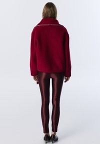 Červený vlněný kabát s vysokým límcem, který má zipový detail na výstřihu. V kombinaci s lesklými vínovými legínami a černými botami.