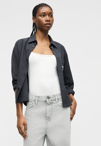 Νεαρή γυναίκα με κοτσίδες, που φοράει λευκό φανελάκι, μαύρο υφασμάτινο πουκάμισο με κουμπιά και ανοιχτό γκρι τζιν, στέκεται μπροστά σε απλό φόντο.