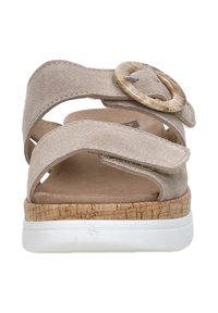 Beige suede sandal with crossover straps and a round, patterned buckle. Cork-textured midsole and white outsole. Flat design.
