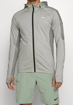 Mand iført lysegrå lynlåstrøje med hætte og meshpaneler samt neonlynlåse, kombineret med lysegrønne Nike-shorts.