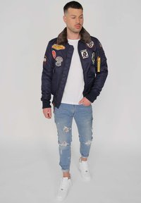 Marineblaues Bomberjackett mit einem Kunstfellkragen, mehreren bunten Patches, Reißverschlusstaschen, gerippten Bündchen, kombiniert mit hellblauen, zerrissenen Jeans und weißen Turnschuhen.