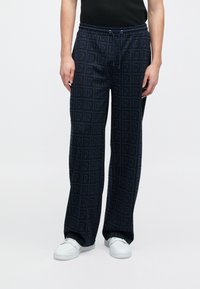 Tumši zili sporta bikses ar visaptverošu logo rakstu, elastīgs jostasvīrs ar auklu, brīva piegriezuma un kombinētas ar baltām kedām.