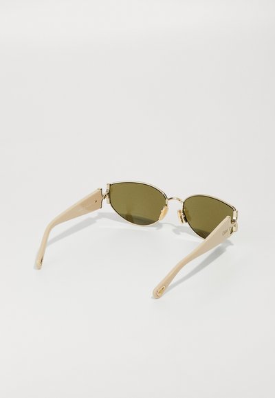 Gafas de sol ovaladas con lentes verdes, acentos metálicos dorados y brazos de plástico beige, que presentan un diseño elegante y una sutil marca en la patilla.