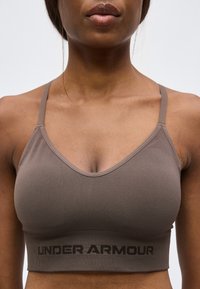 Γυναίκα που φορά καφέ αθλητικό σουτιέν Under Armour με λεπτές τιράντες και εμφανές λογότυπο στο στήθος, εμφανιζόμενη από τους ώμους μέχρι το ανώτερο μέρος της κοιλιάς.
