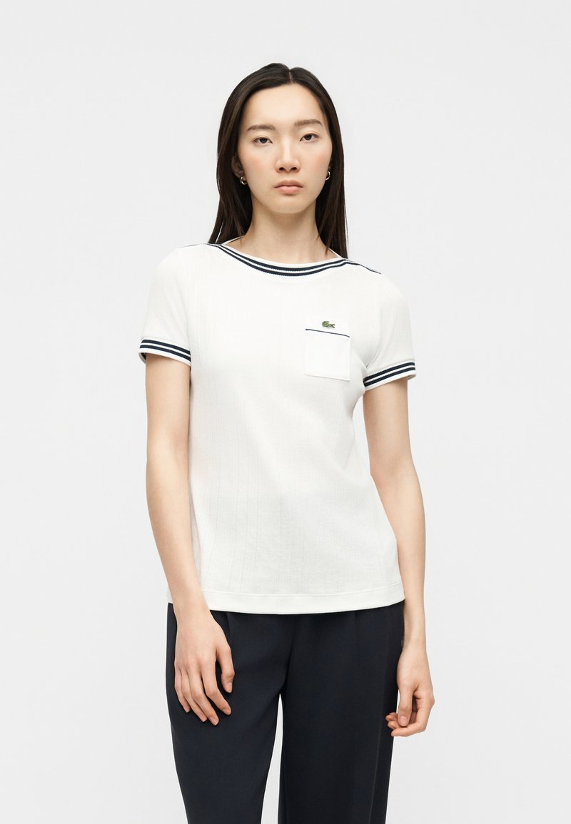Femme portant un haut blanc à manches courtes avec une bordure bleu marine aux manches et au col, pantalon bleu marine, debout devant un fond clair uni.