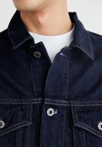 Mörk denimjacka med knäppning fram, två bröstfickor och krage. Har kontrasterande gul sömnad och glänsande metallknappar.