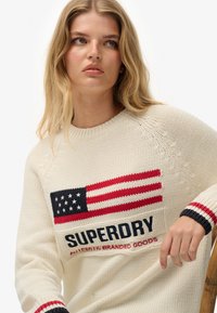 Suéter de punto crema con una gran bandera estadounidense y el texto "SUPERDRY" en azul marino y rojo. Puños acanalados con rayas rojas y azul marino.