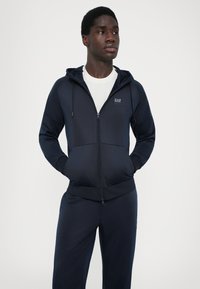 Sweat à fermeture éclair navy avec un design ajusté, comportant une poche avant et une capuche avec cordon de serrage ajustable. Fabriqué en tissu lisse et léger.