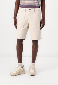 Beige, térdnél közepes hosszúságú short, elülső és oldalsó zsebekkel, világos színű sportcipővel és szürke zoknival párosítva. Az anyaga sima tapintású.