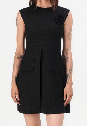 Robe noire texturée sans manches avec poches avant et un détail de rose en tissu sur l'épaule gauche, portée par une personne tatouée.