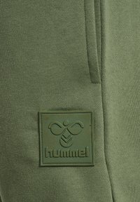 Grønt tekstureret stof med matchende grøn gummipatch-logo, der viser en stiliseret bi og teksten "hummel" nedenfor på tøj.
