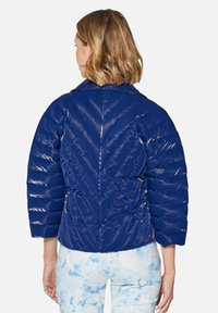 Marineblaue Steppjacke mit glänzender Oberfläche, die horizontale Baffle und ein Zickzackmuster auf dem Rücken aufweist. Abgeschnittene Ärmel.